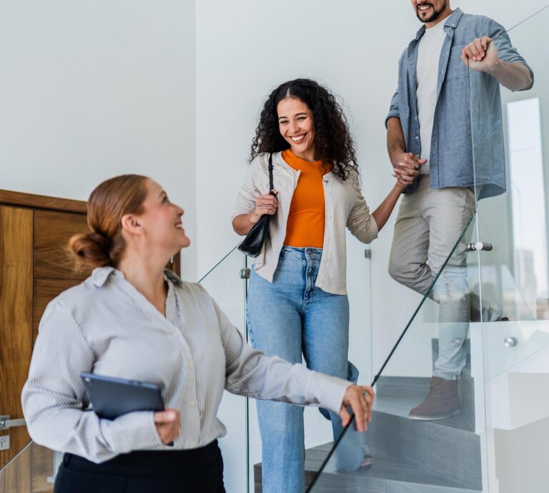Eine Maklerin zeigt einer Frau und einem Mann eine Wohnung. Alle gehen hintereinander die Treppe runter.  | © Getty Images