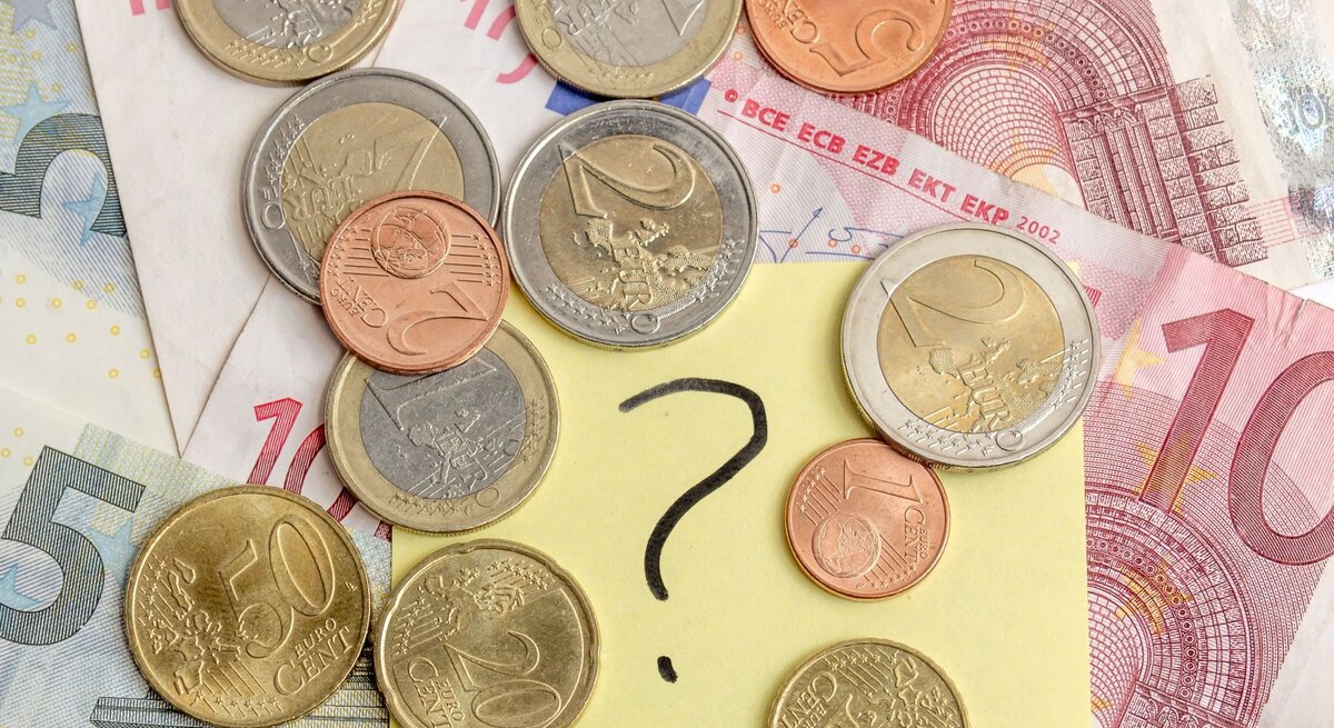 Verschiedene Euro-Münzen und -Scheine liegen auf einem Tisch, in der Mitte ein gelber Zettel mit einem großen Fragezeichen.