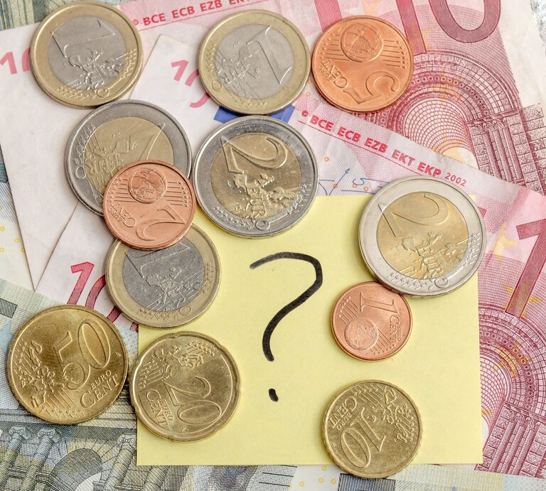 Verschiedene Euro-Münzen und -Scheine liegen auf einem Tisch, in der Mitte ein gelber Zettel mit einem großen Fragezeichen. | © Adobe Stock/simontk