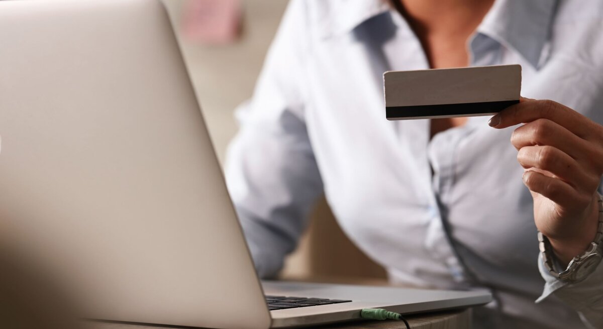 Zu sehen ist eine Frau, die eine Kreditkarte hält und am Laptop sitzt.