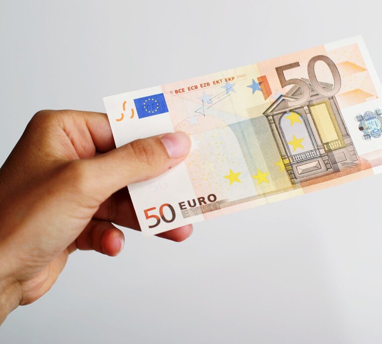 Ein 50-Euro-Schein wird von einer Hand mit leicht gebogenen Fingern gehalten, vor einem schlichten weißen Hintergrund. | © Adobe Stock/Magnus
