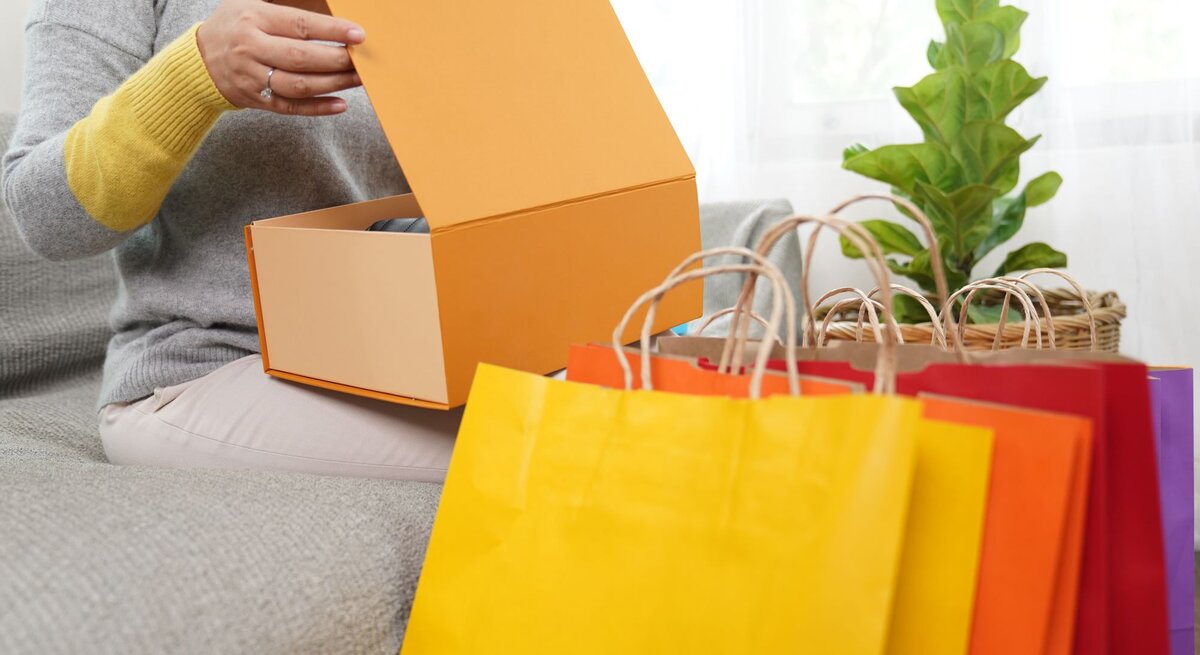 Eine Frau öffnet ein Paket und bunte Einkaufstüten stehen zu ihren Füßen.