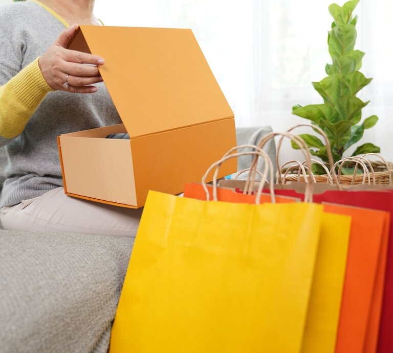 Eine Frau öffnet ein Paket und bunte Einkaufstüten stehen zu ihren Füßen. | © gettyimages/ wera Rodsawang