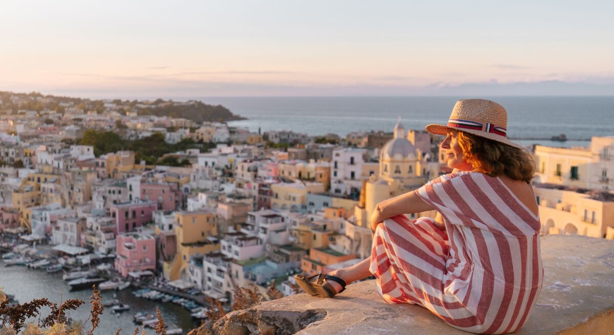 Zu sehen ist eine Frau mittleren Alters, die auf einer Mauer sitzt und auf eine mediterrane Stadt hinunterguckt.