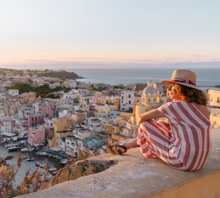 Zu sehen ist eine Frau mittleren Alters, die auf einer Mauer sitzt und auf eine mediterrane Stadt hinunterguckt. | © GettyImages/Mario Martinez