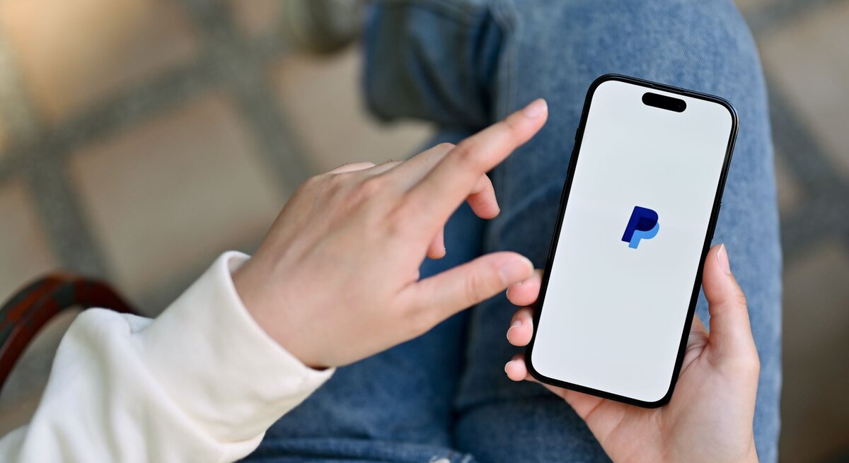 Eine Person in Jeans und weißem Pullover hält ein Smartphone mit dem PayPal-Logo auf dem Bildschirm und tippt mit einer Hand darauf.