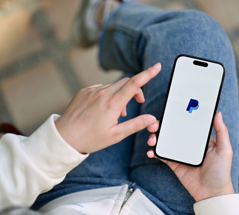 Eine Person in Jeans und weißem Pullover hält ein Smartphone mit dem PayPal-Logo auf dem Bildschirm und tippt mit einer Hand darauf. | © Adobe Sttock/bongkarn