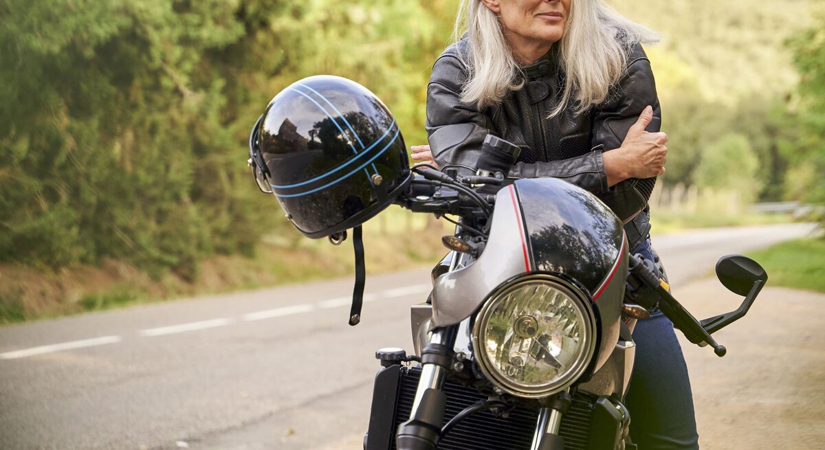 Zu sehen ist eine Frau mittleren Alters mit weißen Haaren, die sich lässig über ihr Motorrad lehnt
