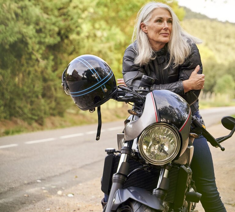 Zu sehen ist eine Frau mittleren Alters mit weißen Haaren, die sich lässig über ihr Motorrad lehnt | © GettyImages/VEAM Visuals