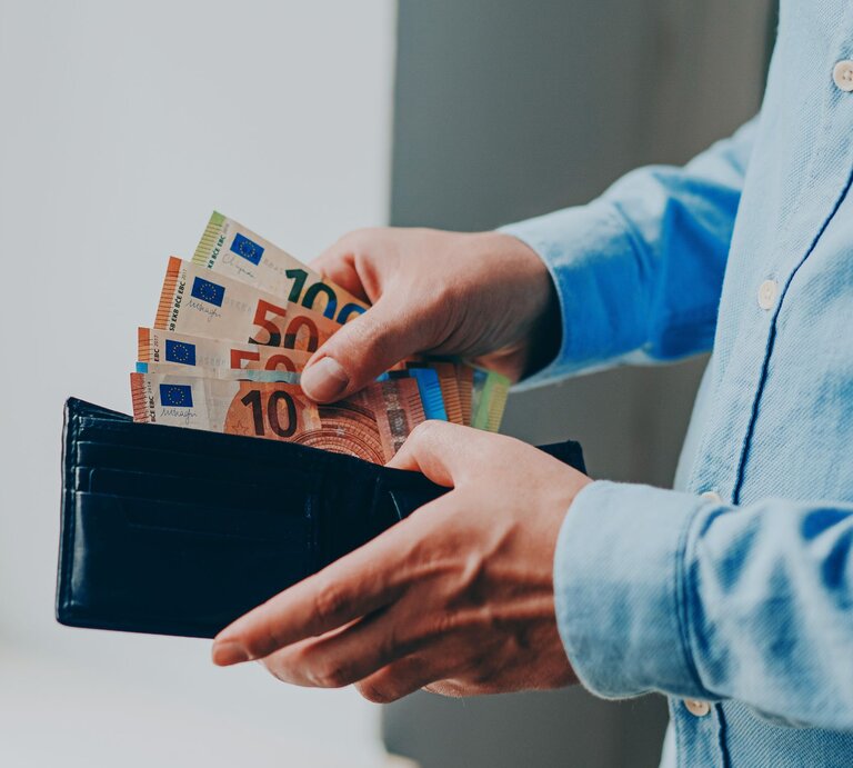 Zu sehen ist eine Hand, die Geld in ein Portemonnaie steckt. | © GettyImages/Olena Malik