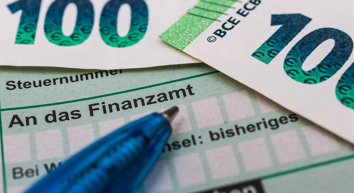 Zwei 100-Euro-Scheine liegen auf einem Steuerformular mit der Aufschrift "An das Finanzamt", daneben ein blauer Kugelschreiber.