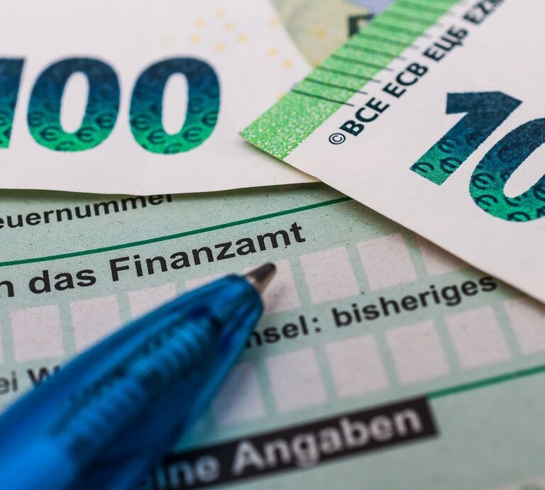 Zwei 100-Euro-Scheine liegen auf einem Steuerformular mit der Aufschrift "An das Finanzamt", daneben ein blauer Kugelschreiber. | © Adobe Stock/Stockfotos-MG