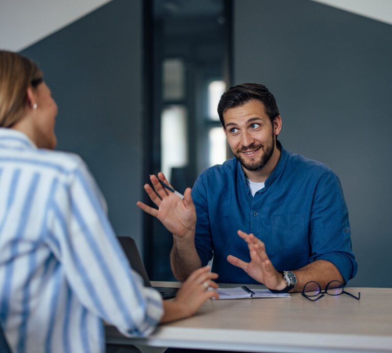 Ein junger Mann mit Bart und blauer Bluse gestikuliert freundlich in einem Gespräch mit einer blondhaarigen Frau in gestreifter Bluse. | © Adobe Stock/bnenin