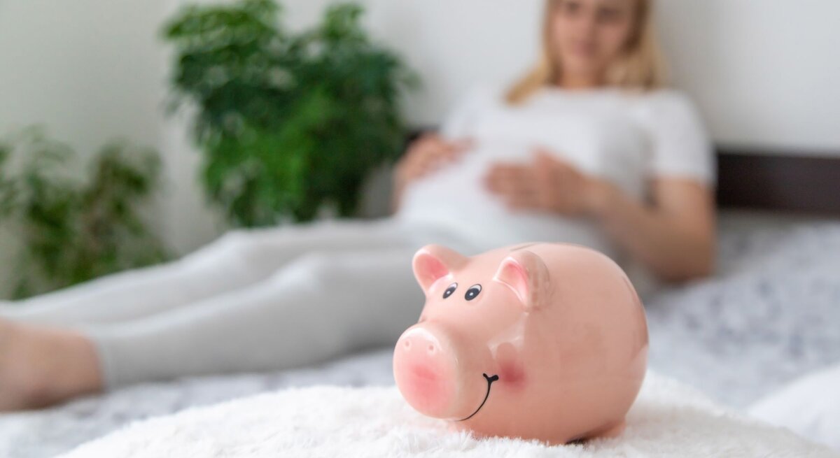 Ein rosa Sparschwein mit lachendem Gesicht steht auf einer flauschigen weißen Decke, im Hintergrund liegt eine schwangere Frau in Weiß.