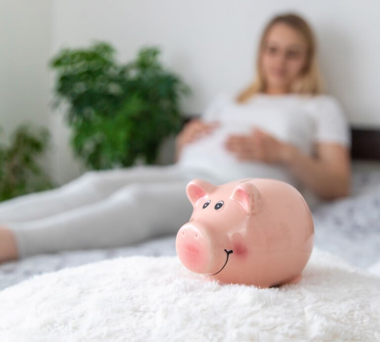 Ein rosa Sparschwein mit lachendem Gesicht steht auf einer flauschigen weißen Decke, im Hintergrund liegt eine schwangere Frau in Weiß. | © Adobe Stock/Anna
