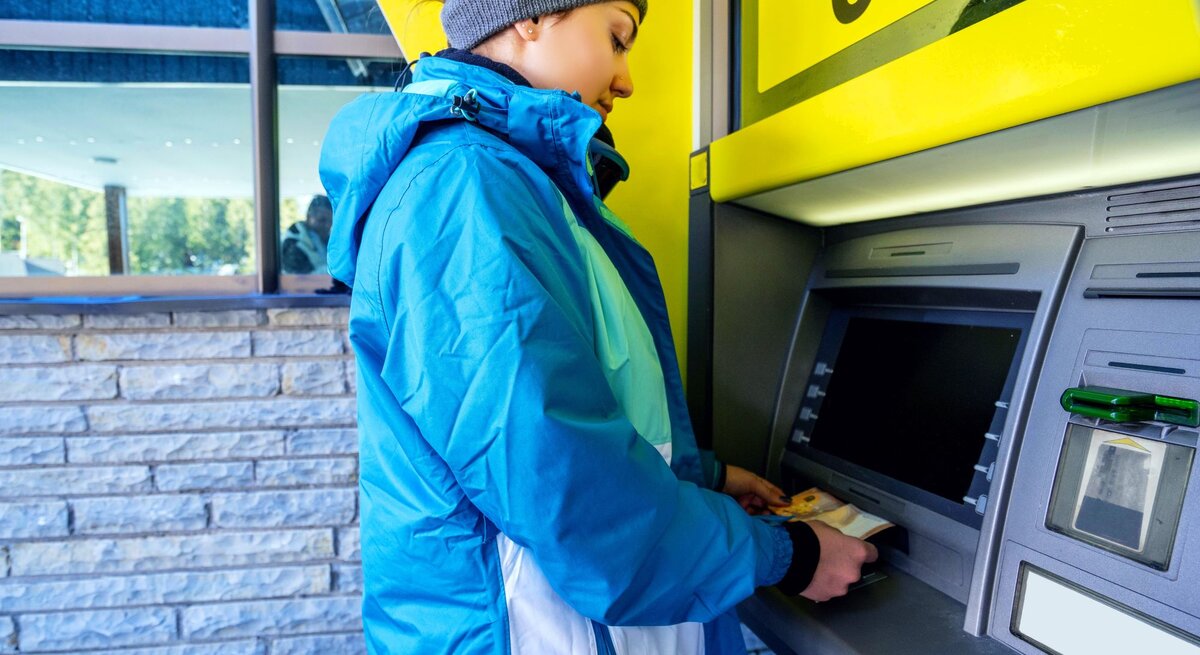 Eine junge Frau in blauer Winterjacke und Stirnband hebt an einem Geldautomaten Geld ab, während sich ihr Gesicht konzentriert nach unten neigt.