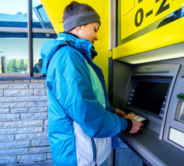 Eine junge Frau in blauer Winterjacke und Stirnband hebt an einem Geldautomaten Geld ab, während sich ihr Gesicht konzentriert nach unten neigt. | © Adobe Stock/alaphotography