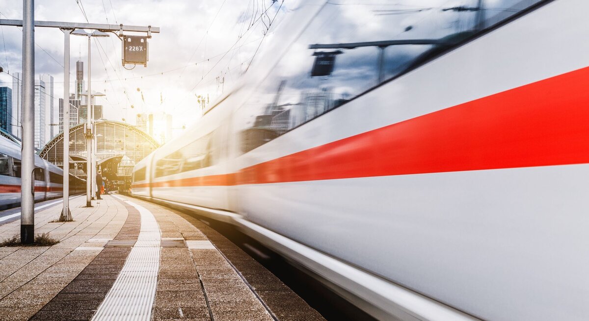 Ein ICE-Zug rauscht mit hoher Geschwindigkeit an einem Bahnsteig vorbei, während die Sonne hinter einer modernen Bahnhofshalle aufgeht.