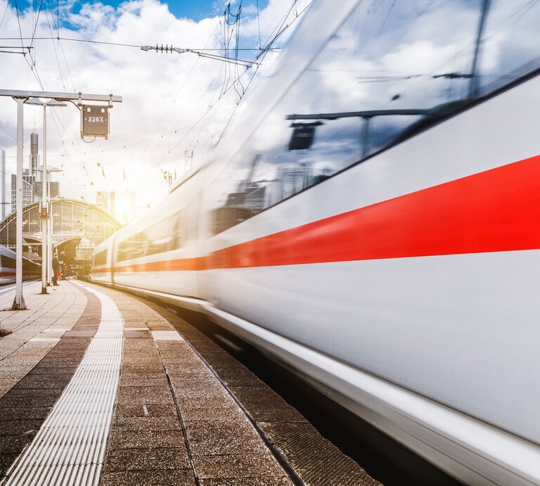 Ein ICE-Zug rauscht mit hoher Geschwindigkeit an einem Bahnsteig vorbei, während die Sonne hinter einer modernen Bahnhofshalle aufgeht. | © Adobe Stock/nokturnal