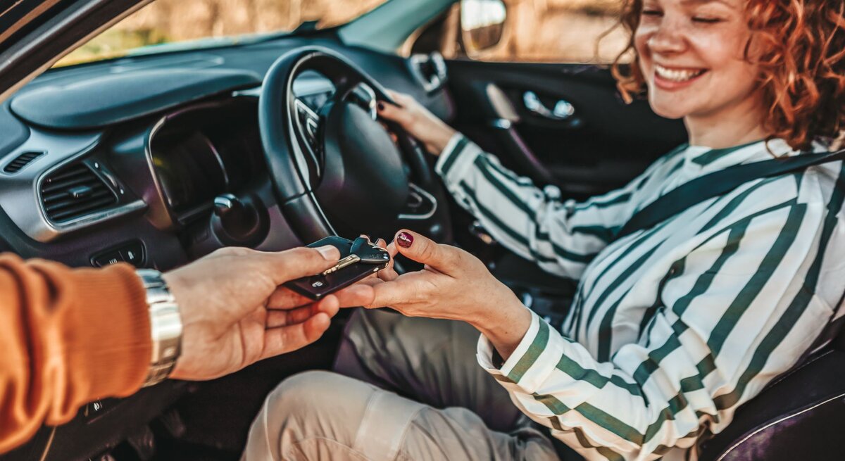 Frau sitzt lächeln im Auto, geöffnete Tür und bekommt den Schlüssel gereicht.