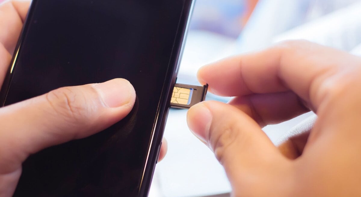 Eine Hand setzt eine goldene SIM-Karte in den geöffneten Kartenslot eines schwarzen Smartphones ein, im Hintergrund ein heller Schreibtisch.