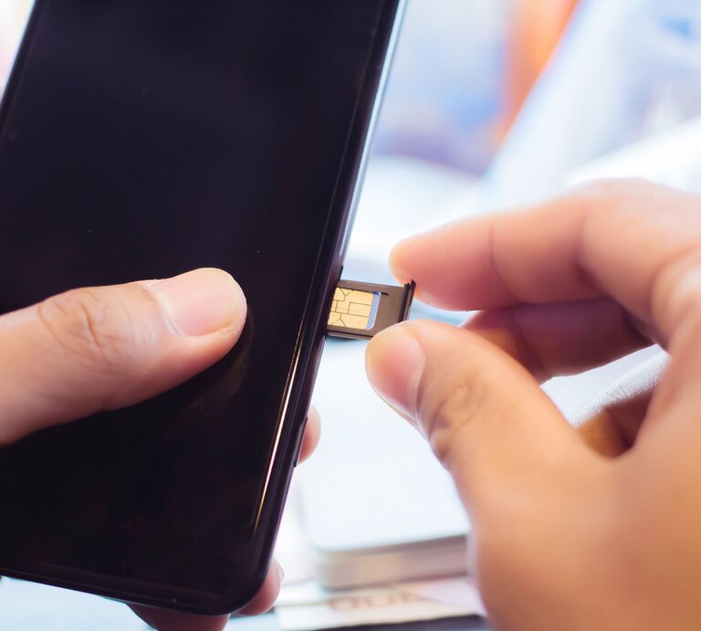 Eine Hand setzt eine goldene SIM-Karte in den geöffneten Kartenslot eines schwarzen Smartphones ein, im Hintergrund ein heller Schreibtisch. | © Adobe Stock/Stock.Foto.Touch