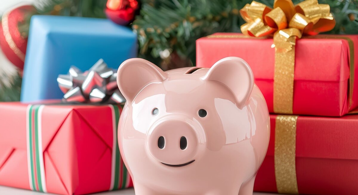 Ein rosa Sparschwein steht vor rot-goldenen Geschenken unter einem geschmückten Weihnachtsbaum mit Kugeln und Zapfen.