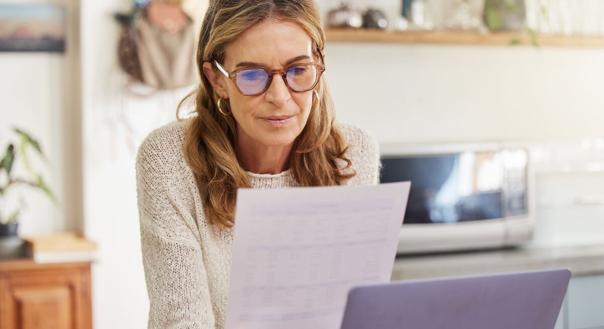 Frau, die konzentriert ein Dokument liest, während sie an einem Laptop sitzt. Sie trägt eine Brille und ist in einer gemütlichen Wohnküche. Im Hintergrund sind Mikrowelle und Pflanzen zu sehen.