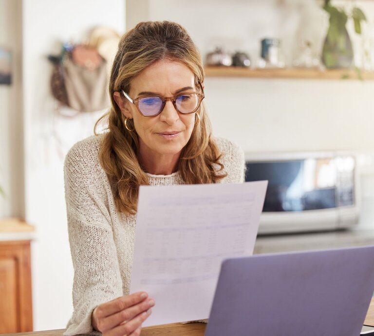 Frau, die konzentriert ein Dokument liest, während sie an einem Laptop sitzt. Sie trägt eine Brille und ist in einer gemütlichen Wohnküche. Im Hintergrund sind Mikrowelle und Pflanzen zu sehen. | © Adobe Stock / N Lawrenson/peopleimages.com