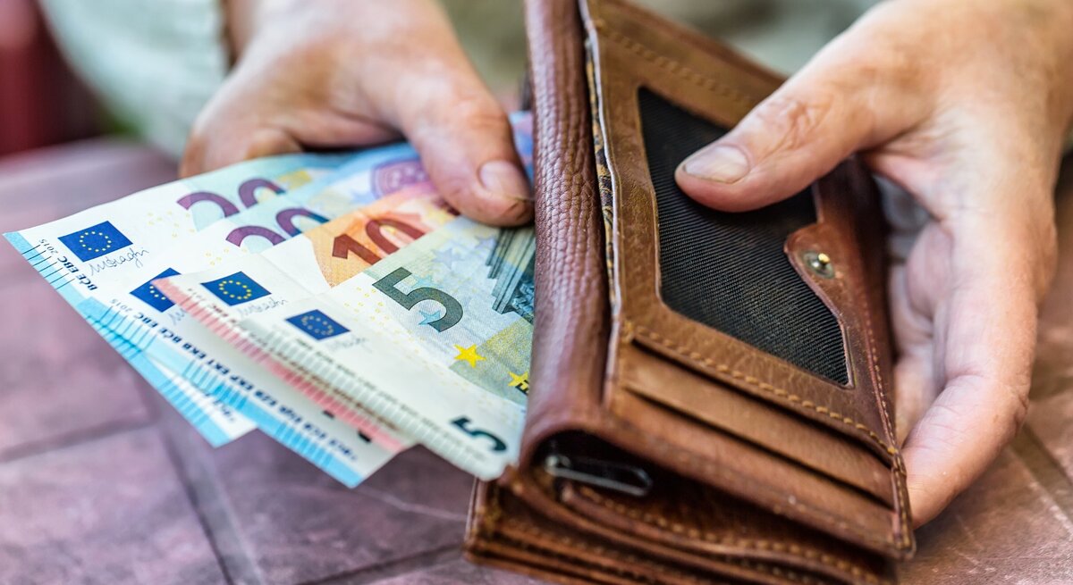 Eine Nahaufnahme von zwei Händen, die ein braunes Lederportemonnaie halten, aus dem mehrere Euro-Banknoten herausragen, darunter Scheine mit den Werten 5, 10, 20 und 50 Euro. Die Szene wirkt, als ob jemand gerade dabei ist, Geld zu zählen oder zu organisieren.