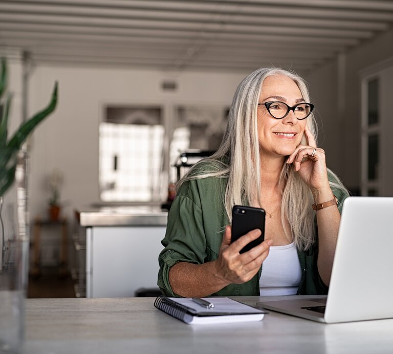 Dame höheren Alters sitzt vor dem PC mit dem Handy in der Hand | © iStock / Ridofranz