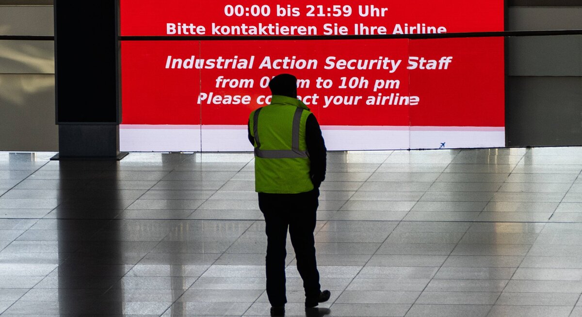Ein Sicherheitsmitarbeiter steht vor eine Streik-Infowand in einem Flughafen.