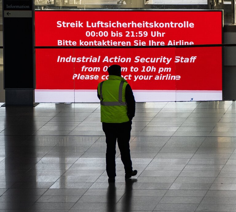 Ein Sicherheitsmitarbeiter steht vor eine Streik-Infowand in einem Flughafen. | © IMAGO / Chris Emil Janßen