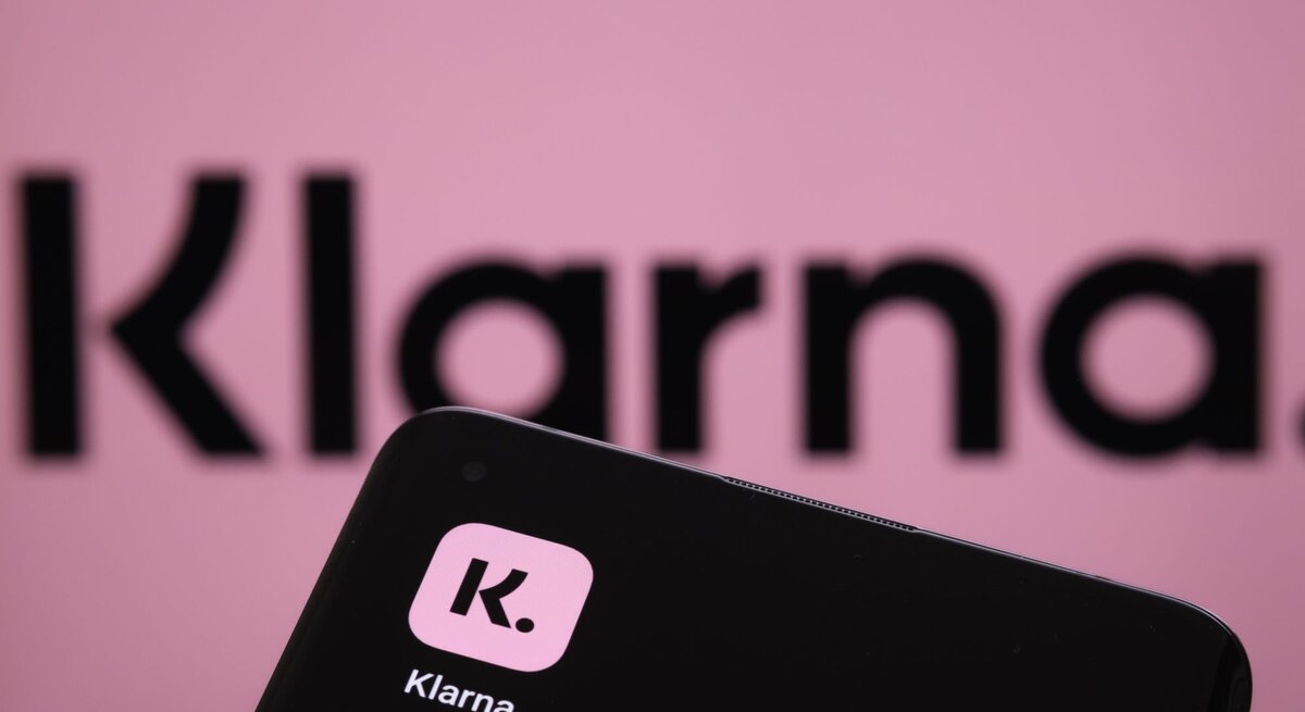 Im Vordergrund ist ein Smartphone mit dem Logo von Klarna, einer bekannten Zahlungs- und Shopping-App, zu sehen. Der Hintergrund zeigt unscharf das Klarna-Logo in Großbuchstaben auf einem rosa Hintergrund. Der Fokus liegt auf dem Smartphone