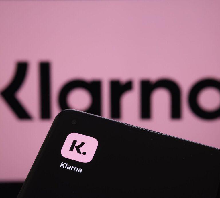Im Vordergrund ist ein Smartphone mit dem Logo von Klarna, einer bekannten Zahlungs- und Shopping-App, zu sehen. Der Hintergrund zeigt unscharf das Klarna-Logo in Großbuchstaben auf einem rosa Hintergrund. Der Fokus liegt auf dem Smartphone | © Adobe Stock/ Ascannio