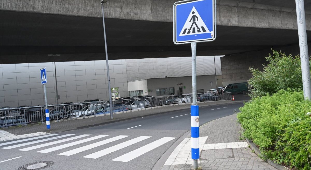 Ein Foto von einem städtischen Straßenbereich mit einem Fußgängerüberweg, erkennbar an den typischen weißen Streifen auf dem Asphalt. Direkt vor dem Zebrastreifen steht ein blau-weißes Verkehrsschild, das auf den Fußgängerüberweg hinweist. Die Umgebung wirkt urban mit einem Betonbauwerk im Hintergrund, unter dem die Straße hindurchführt. Am Straßenrand befindet sich eine Reihe geparkter Autos und auf der rechten Seite ist eine kleine Grünfläche mit Büschen zu sehen