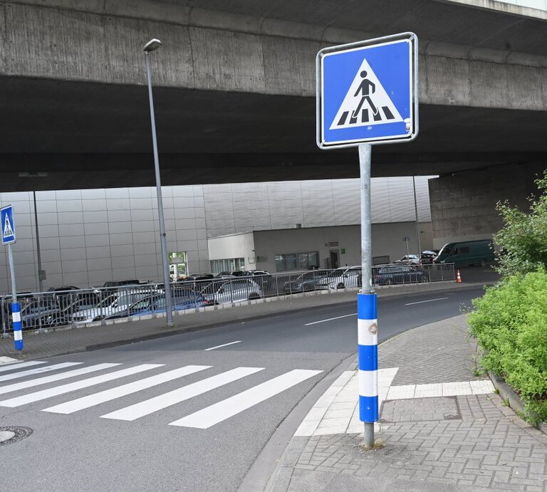 Ein Foto von einem städtischen Straßenbereich mit einem Fußgängerüberweg, erkennbar an den typischen weißen Streifen auf dem Asphalt. Direkt vor dem Zebrastreifen steht ein blau-weißes Verkehrsschild, das auf den Fußgängerüberweg hinweist. Die Umgebung wirkt urban mit einem Betonbauwerk im Hintergrund, unter dem die Straße hindurchführt. Am Straßenrand befindet sich eine Reihe geparkter Autos und auf der rechten Seite ist eine kleine Grünfläche mit Büschen zu sehen | © IMAGO / Horst Galuschka