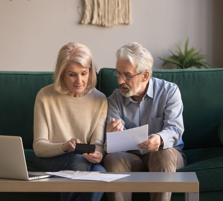 Ein älteres Paar sitzt auf einem grünen Sofa und bespricht konzentriert Dokumente, neben ihnen ein Laptop auf einem Holztisch. | © Adobe Stock / fizkes