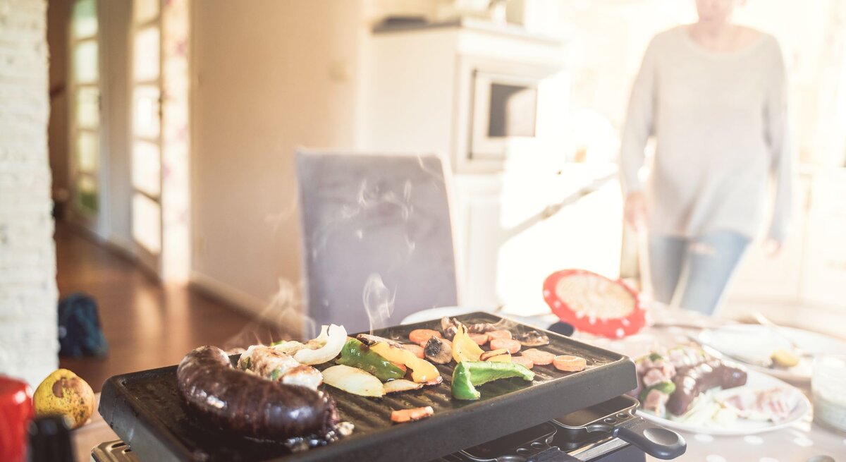 Ein Raclette mit rauchendem Gemüse und Würstchen steht auf einem gedeckten Tisch, im Hintergrund läuft eine Frau durch die Küche.