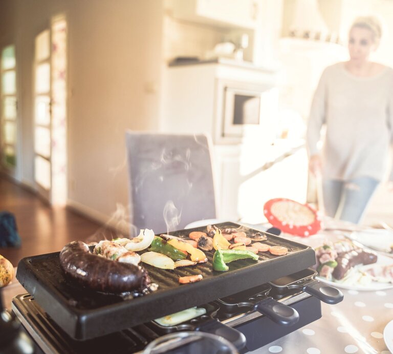 Ein Raclette mit rauchendem Gemüse und Würstchen steht auf einem gedeckten Tisch, im Hintergrund läuft eine Frau durch die Küche. | © Shutterstock/kmfdm1