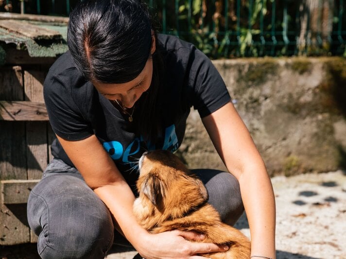 Eine dunkelhaarige Frau in schwarzem Shirt kniet vor einer Hundehütte und streichelt liebevoll einen kleinen braunen Hund. | © PETA
