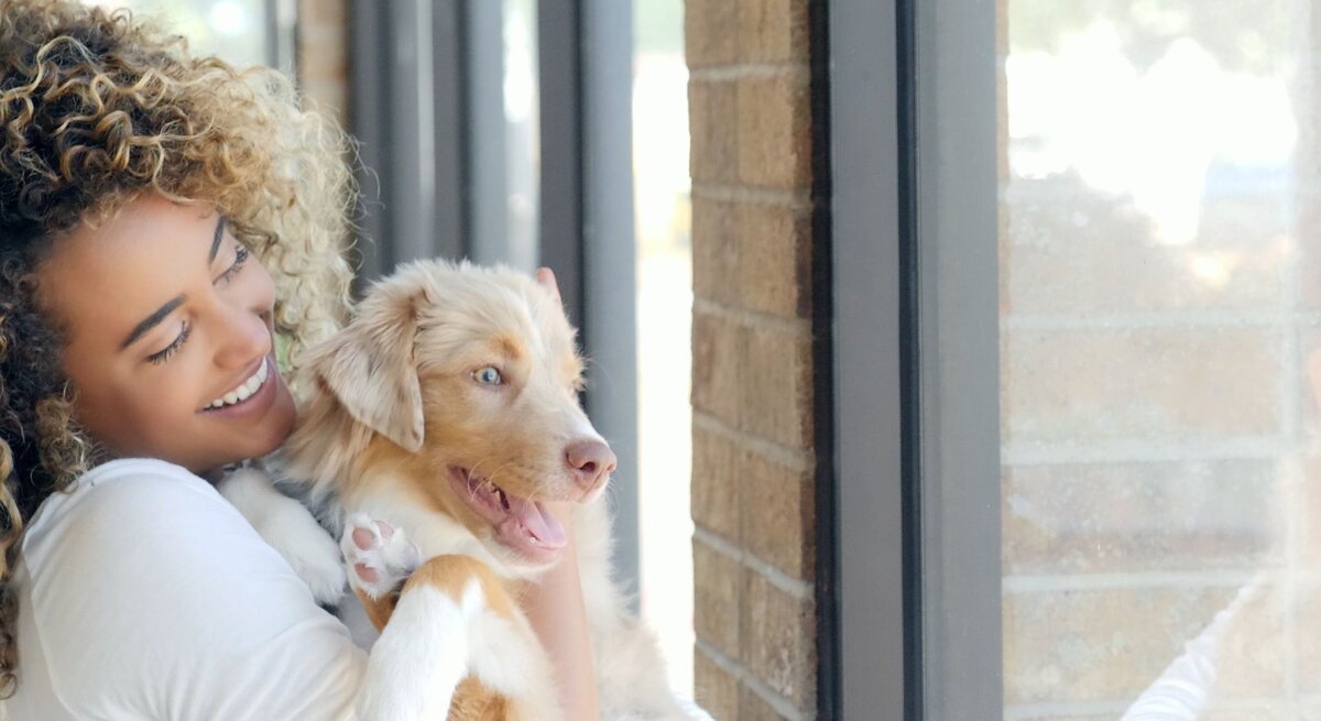 Eine junge Frau mit lockigem Haar hält lächelnd einen hellbraunen, jungen Hund vor einem Fenster.