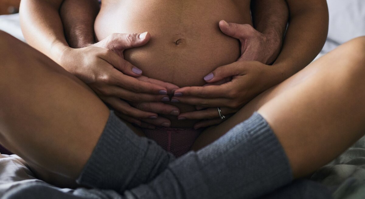 Junge schwangere Frau in rosa Unterwäsche sitzt auf Bett, Partner umarmt sie liebevoll, beide Hände auf Babybauch.