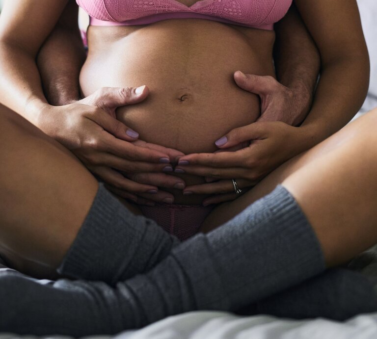 Junge schwangere Frau in rosa Unterwäsche sitzt auf Bett, Partner umarmt sie liebevoll, beide Hände auf Babybauch. | © Adobe Stock / CamPen2000/peopleimages.com