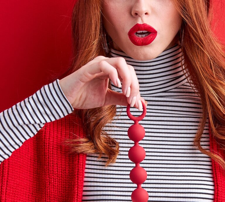 Junge Frau mit roten Lippen und langen roten Haaren hält rote Liebeskugeln vor rot gestreiftem Hintergrund in Brusthöhe. | © Adobe Stock / 