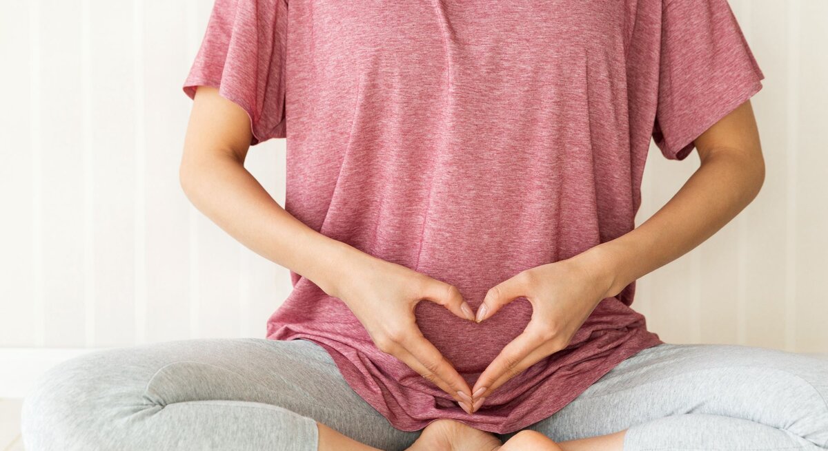 Junge Frau in rosa Shirt und grauer Leggings sitzt im Schneidersitz und formt mit den Händen ein Herz über ihrem Unterbauch.
