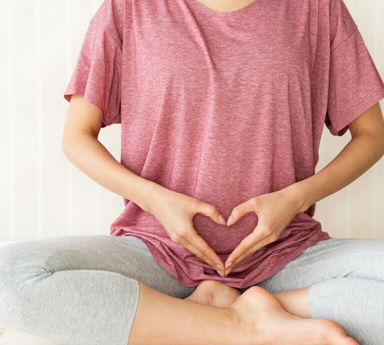Junge Frau in rosa Shirt und grauer Leggings sitzt im Schneidersitz und formt mit den Händen ein Herz über ihrem Unterbauch. | © Adobe Stock / kei907