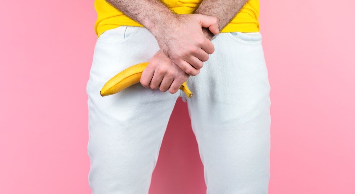 Junger Mann in gelbem T-Shirt hält eine Banane provokant vor seinem Schritt, vor rosa Hintergrund und weißer Hose.