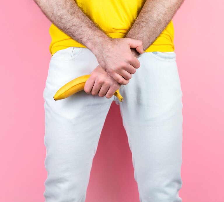 Junger Mann in gelbem T-Shirt hält eine Banane provokant vor seinem Schritt, vor rosa Hintergrund und weißer Hose. | ©  Getty Images / Ildar Abulkhanov