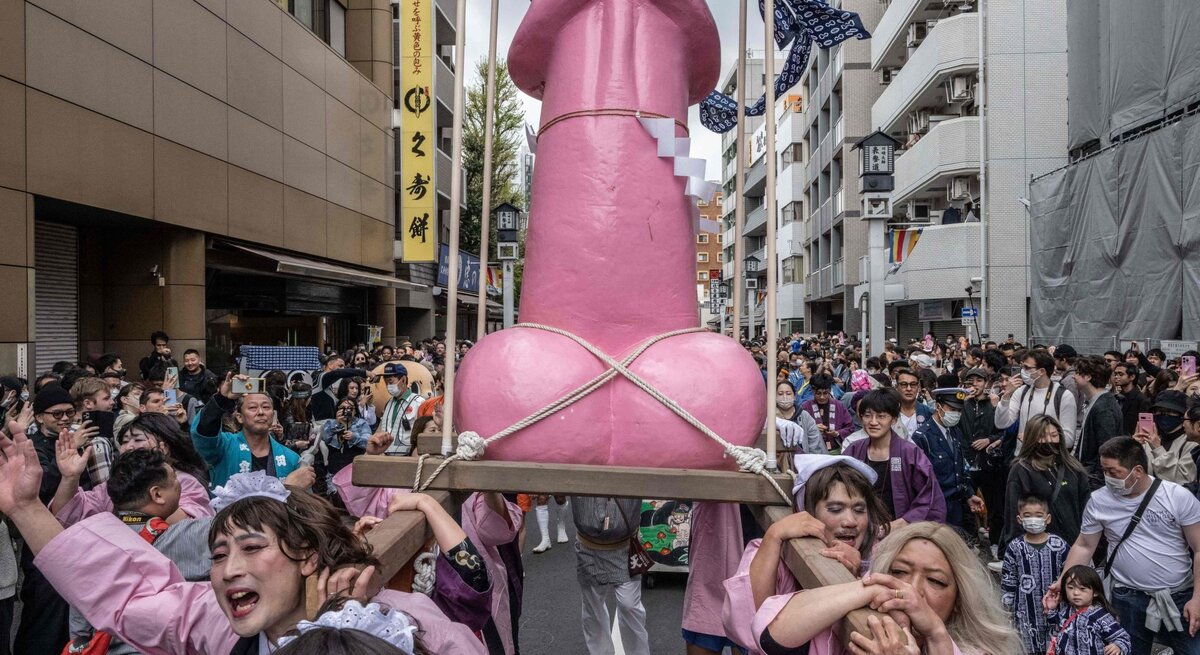 Menschen in bunten Kostümen tragen bei einem Straßenfest in Japan eine große pinke Penis-Skulptur durch eine dicht gedrängte Menge.